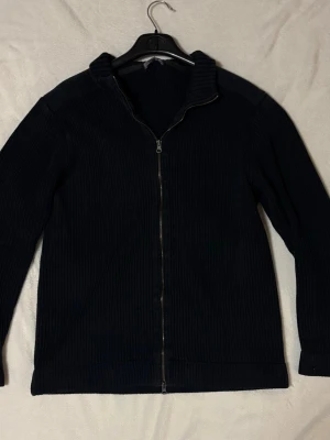 Mörkblå ribbad ziptröja - En stilren mörkblå ziptröja med ribbad struktur och hög krage. Tröjan har hel dragkedja framtill och långa ärmar, perfekt för lager på lager. Passar dig som gillar en enkel men snygg look.