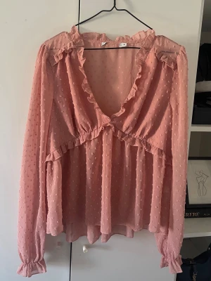 Rosa volangblus med prickig struktur - Söt rosa blus med volangdetaljer och djup V-ringning. Blusen har långärmade ärmar med resår vid ärmslut och är gjord i ett tunt, prickigt tyg som ger en luftig känsla. 