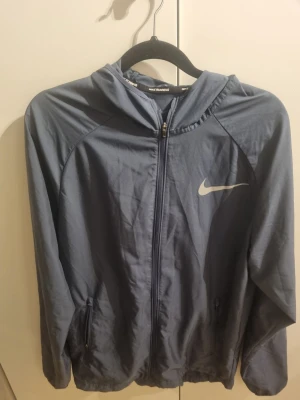 Blå Nike vindjacka med huva - Säljer en blå vindjacka från Nike med huva och dragkedja framtill. Jackan har två fickor med dragkedja och Nike-logga på bröstet. Perfekt för löpning och sportiga aktiviteter, lätt material som andas.