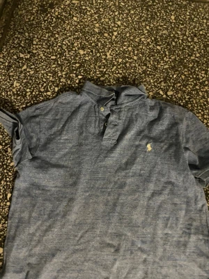 Blå kortärmad henley från Polo Ralph Lauren - Snygg blå kortärmad henley från Polo Ralph Lauren med klassisk knappslå och den ikoniska broderade loggan på bröstet. Skjortan är gjord i mjuk bomull och har en avslappnad passform som passar perfekt till jeans eller shorts.