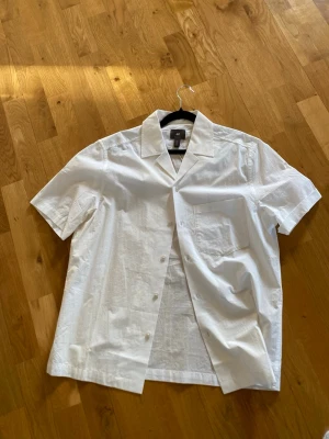 Vit kortärmad skjorta från H&M - Fräsch vit skjorta från H&M i regular fit med korta ärmar och klassisk krage. Skjortan har knäppning framtill och en bröstficka. Perfekt för en stilren och enkel look. Tillverkad i ett lätt och svalt material. Lappen kvar