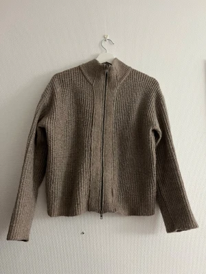 Beige stickad ziptröja - Stickad tröja i beige nyans med hel dragkedja framtill och hög krage. Tröjan har ribbstickad struktur och långa ärmar. Perfekt för lager-på-lager och chill dagar. Materialet ser ut att vara ull eller ullmix.