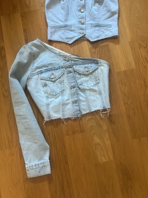 Asymmetrisk ljusblå jeansjacka - Trendig ljusblå jeansjacka från NA-KD med asymmetrisk design och rå, fransad nederkant. Jackan har en ärm, två bröstfickor med knappar och klassiska jeansknappar framtill. Perfekt för dig som vill sticka ut med en unik look.
