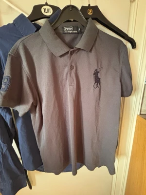 Grå pikétröja från Polo Ralph Lauren - Snygg grå pikétröja från Polo Ralph Lauren med klassisk krage och knäppning framtill. Tröjan har broderad logga på bröstet och en siffra på ärmen. Perfekt till jeans eller shorts för en clean look.