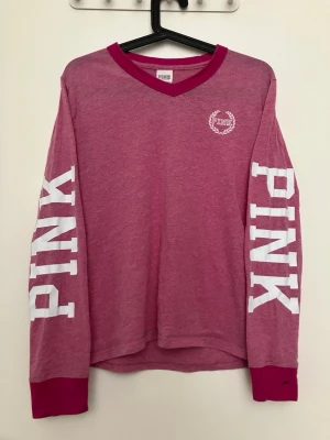 Rosa långärmad topp från PINK - Långärmad topp från PINK i en härlig rosa nyans med vita tryck på båda ärmarna och liten logga på bröstet. V-ringad med mörkare rosa kant och mjukt bomullsmaterial. Perfekt för en avslappnad och cool stil.