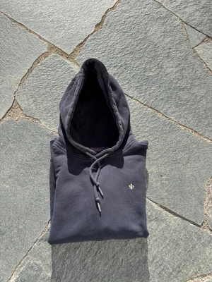 Svart hoodie från Morris - Snygg svart hoodie från Morris med klassisk huva och dragsko. Liten broderad logga på bröstet och mjukt material som känns skönt mot huden. Perfekt för en avslappnad och stilren look.
