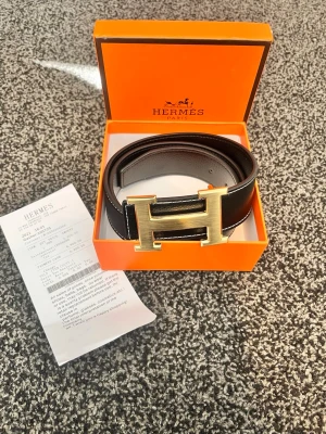 Svart H skärp med guldfärgat H-spänne - Stilrent svart skärp från H@rmes med ikoniskt H-spänne i guldfärgad metall. Skärpet är gjort i exklusivt skinn och levereras i originalask. Perfekt accessoar för att lyfta din outfit med en lyxig touch.