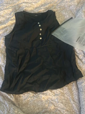 Svart ärmlös linnetopp med knappar - Svart ärmlös linnetopp med rund halsringning och fem vita knappar framtill. Enkel och stilren design som passar till många olika outfits. Aldrig använd