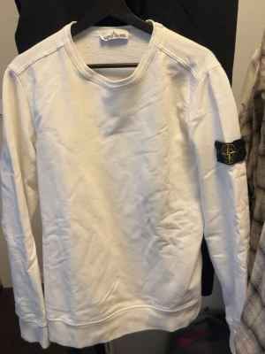 Vit sweatshirt från Stone Island ( ÄKTA) - Vit sweatshirt från Stone Island med klassisk rund hals och ribbade muddar. Tröjan har den ikoniska svarta logotypen med kompass på vänster ärm. Tillverkad i mjukt bomullsmaterial som är skönt mot huden. Perfekt för en stilren och avslappnad look, Sitter lite tajt och mig som är 180 lång!!. För att authenticate checka den skriv till mig!!