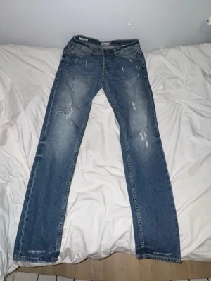 Jeans med slitningar - Säljer ett par blå jeans med raka ben och slitna detaljer på låren och knäna. Jeansen har klassisk femficksdesign och är gjorda i denim. Perfekta för en avslappnad och trendig look.