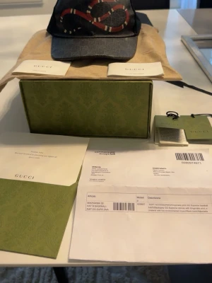 Gucci keps - Snygg svart keps från Gucci med ikoniskt GG-mönster och en rödvit orm broderad framtill. Kepsen har böjd skärm och justerbar baksida i mesh. Materialet är canvas och mesh, vilket ger en cool och lyxig känsla. Perfekt för dig som vill sticka ut med en designeraccessoar.