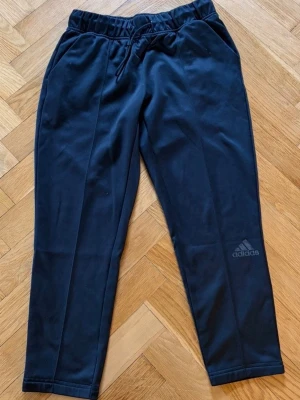 Svarta mjukisbyxor från Adidas - Svarta mjukisbyxor från Adidas med dragsko i midjan och två sidofickor. Byxorna har en smalare passform och Adidas-logga på ena benet. Perfekta för chill eller träning, tillverkade i mjukt material.