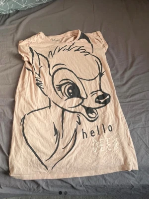 Beige pyjamas med Bambi-tryck - Supersöt beige pyjamas med stort Bambi-tryck och texten 'hello deer' framtill. T-shirten har korta ärmar och är gjord i mjuk bomull, perfekt för mysiga kvällar. Avslappnad passform och rund halsringning. Det är ett nattlinne