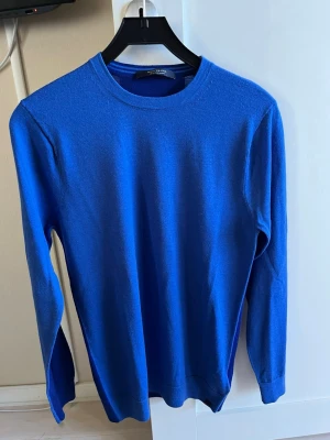 Blå stickad tröja Scotch & Soda - En stilren blå stickad tröja från Scotch & Soda med rund halsringning och långa ärmar. Tröjan har en enkel och klassisk design, perfekt att matcha med jeans eller chinos. Materialet känns mjukt och lätt, vilket gör den skön att bära. Finns ett litet mindre hål de bild 3, syns knappt och lätt och laga. Därav lite lägre pris.