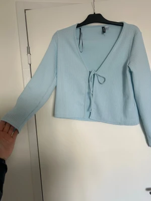Ljusblå ribbad kofta från H&M Divided - Ljusblå ribbad kofta från H&M Divided med knytband framtill och v-ringad hals. Koftan har långa ärmar och är tillverkad i ett mjukt, stretchigt material som ger en skön passform. Perfekt att slänga över en topp för en chill look.