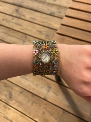 Blommig vintageklocka med bred länk, oklart om batteriet behövs bytas - Unik och färgglad klocka med bred metallarmband dekorerat med små blommor i olika färger. Urtavlan är vit med svarta siffror och omges av detaljerade snirkliga mönster. Perfekt för dig som gillar retro och boho vibes.