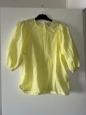 Gul blus med puffärm från Line of Oslo - Säljer en ljusgul blus från Line of Oslo med rund halsringning och puffiga trekvartsärmar. Materialet är lätt och lite transparent, vilket ger en luftig känsla. Perfekt för dig som gillar färg och vill ha något som sticker ut.