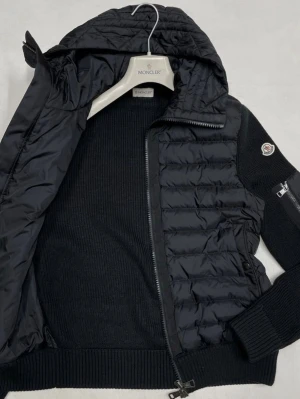 Svart cardiganjacka från Moncler - Snygg svart cardiganjacka från Moncler med quiltad framsida och stickade ärmar. Jackan har dragkedja, huva och Moncler-logga på ärmen. Materialmix med dunfylld front och ribbstickade muddar ger en modern look.
