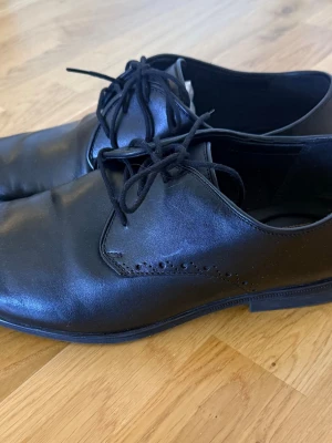 Svarta läderskor från Clarks - Stilrena svarta läderskor från Clarks med klassisk snörning och diskreta perforerade detaljer vid sidan. Skorna har en elegant, något spetsig tå och låg sula, perfekta för en dressad look. Helt nya!