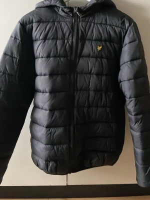 Svart pufferjacka från Lyle & Scott - Snygg svart pufferjacka från Lyle & Scott Junior med huva och dragkedja framtill. Jackan har det klassiska Lyle & Scott-logotypen i gult på bröstet och quiltad design som håller värmen. Perfekt för kalla vinterdagar och kyliga kvällar. Bara att höra av dig om du är intresserad!
