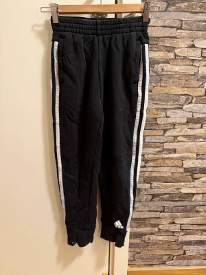 Svarta Adidas mjukisbyxor med vita ränder - Säljer ett par svarta mjukisbyxor från Adidas med klassiska vita ränder längs sidorna och logga vid benet. Byxorna har resår i midjan och vid bensluten samt sidofickor. Perfekta för chill eller träning.