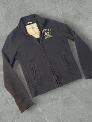 Mörkblå zip-hoodie Abercrombie & Fitch | XL - Mörkblå hoodie med dragkedja från Abercrombie & Fitch. Broderad logga och text '92 New York' på bröstet. Mjuk insida och två fickor framtill. Perfekt för en avslappnad och sportig stil.