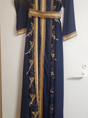 Marinblå långklänning (kaftan) med guldiga broderier - Elegant marinblå (kaftan) långklänning med vackra guldiga broderier och detaljer längs ärmar, midja och framtill. Klänningen har lång ärm, markerad midja med bälte och ett klassiskt, flytande fall. Perfekt för dig som vill sticka ut med stilrena detaljer och lyxig känsla.
