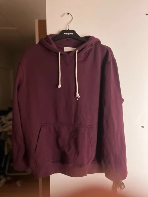 Vinröd hoodie från Pier One - Säljer en vinröd hoodie från Pier One med klassisk känguruficka och vita dragsnören i huvan. Hoodien har en avslappnad passform och är tillverkad i mjukt bomullsmaterial. Perfekt för dig som gillar en enkel och stilren look.