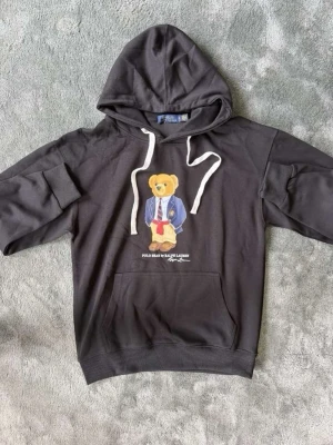 Svart hoodie Polo Bear Ralph Lauren - Säljer en svart hoodie från Ralph Lauren med den ikoniska Polo Bear-trycket framtill. Hoodien har vita dragsnören, stor känguruficka och ribbade muddar. Materialet är mjukt och bekvämt, perfekt för en avslappnad stil.