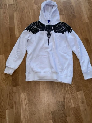 Vit hoodie med svarta vingar från Marcelo Burlon - Snygg vit hoodie från Marcelo Burlon med ikoniskt svart vingtryck över axlar och bröst, samt på ryggen. Hoodien har huva med dragsko och en stor magficka. Perfekt för dig som vill sticka ut med en unik streetstyle-look.