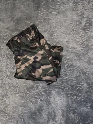  Cargo-byxor Woodland (ART 16) -  Rejäla cargobyxor i klassiskt skogskamouflage. I nyskick med kraftigt tyg och klara färger. Perfekt passform med resår i midjan och rymliga fickor. Inget slitage vid bensluten. • Storlek: innerbernslängd 68cm lång 105 cm midja 89