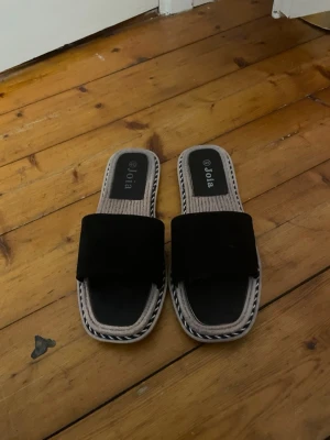 Svarta sandaler från Joia - Snygga svarta sandaler från Joia med bred rem i mockaimitation och fyrkantig tå. Sulan har ett flätat mönster i beige och svart samt kontrastkant. Perfekta för en avslappnad och trendig look. Små i storleken passar 38/39