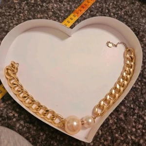 Chunky guldfärgat halsband med pärlor - Säljer ett chunky halsband i guldfärgad metall med stora länkar och två dekorativa pärlor framtill. Mellan pärlorna sitter en liten detalj med glittrande stenar. Perfekt statement piece för dig som gillar att sticka ut.