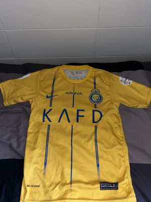 Gul Al Nassr matchtröja från Nike - Säljer en gul Al Nassr matchtröja från Nike med blå detaljer och klubbmärke på bröstet. Tröjan är tillverkad i Dri-FIT material som andas och har korta ärmar. Perfekt för fotbollsträning eller att bära som supporter.