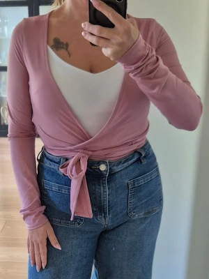 Rosa omlottopp med knytband - Supersnygg rosa omlottopp med långa ärmar och knytband i midjan. Toppen har en djup v-ringning och är i ett mjukt, stretchigt material som sitter skönt på kroppen. Storlek S/M