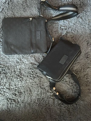 Svarta axelväskor från Gucci - Två svarta axelväskor från Gucci i canvas med diskret GG-mönster och svarta läderdetaljer. Ena väskan har en dragkedja framtill och den andra har dubbla dragkedjor upptill. Båda har justerbara axelremmar med silverfärgade detaljer.