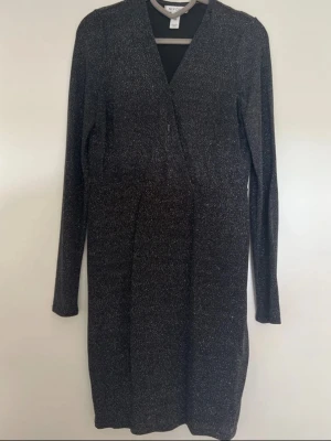 Svart glittrig omlottklänning  - Svart glittrig klänning från New Look med omlott-skärning och lång ärm. Klänningen har en v-ringad hals och är i ett stretchigt material som ger en snygg passform. Perfekt för dig som vill sticka ut med lite extra glitter.