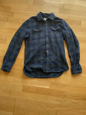 Mörkblå jeansskjorta från Levi's - Mörkblå jeansskjorta från Levi's med klassisk krage, två bröstfickor med lock och knappar framtill. Skjortan är långärmad och har en avslappnad passform. Perfekt för dig som gillar en tidlös och avslappnad stil.