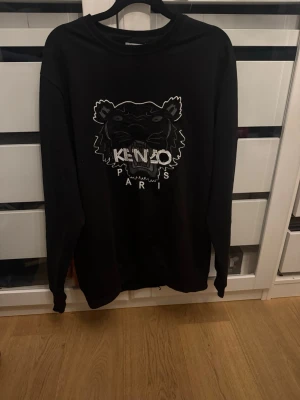 Svart Kenzo hoodie med tigertryck - Svart hoodie från Kenzo med stort broderat tigerhuvud och texten 'KENZO PARIS' på bröstet. Klassisk passform med långa ärmar och ribbade muddar. Perfekt för dig som gillar streetwear och vill sticka ut med en ikonisk design. Passar storlek M-XL.