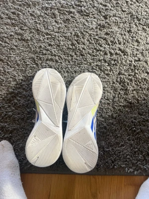 Vita Adidas sneakers med färgglada detaljer - Säljer ett par vita Adidas sneakers med ventilerande mesh och färgglada detaljer i blått, gult och rött. Skorna har en låg profil och sportig design med vita snören och mönstrad sula. Perfekta för dig som gillar stilrena men ändå unika sneakers.