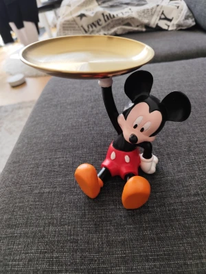 Musse Pigg smyckesfat i plast och metall - Snyggt smyckesfat med Musse Pigg-figur i plast som håller upp ett runt fat i guldfärgad metall. Perfekt för att förvara ringar, örhängen eller andra små accessoarer. Färgerna är svart, röd, vit, orange och guld.