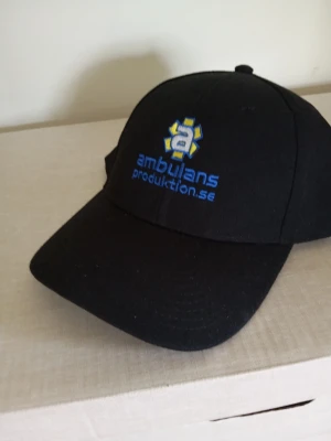 Svart keps Ambulans - Svart keps med böjd skärm och broderad logga från Ambulansproduktion.se i blått och gult framtill. Klassisk modell med sex paneler och justerbar baksida. Perfekt för dig som gillar unik streetwear med tydlig profil.