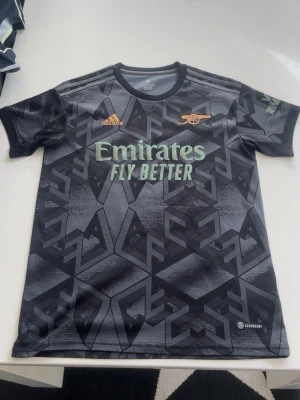 Arsenal bortatröja Adidas svart/grå - Snygg Arsenal bortatröja från Adidas i svart och grått med geometriskt mönster. Tröjan har klubbens logga i guld, Adidas-logga och sponsortryck framtill. Tillverkad i lätt och ventilerande material, perfekt för fotbollsträning eller match.