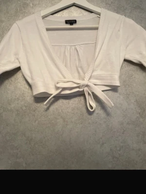 Vit kort omlottkofta - Söt vit kort omlottkofta med knytband framtill och ribbade kanter. Perfekt att slänga över en topp eller klänning för en trendig lager-på-lager-look. Koftan har korta ärmar och en mjuk, stretchig känsla.