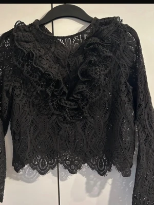Svart spetsblus med volangdetaljer - Trendig svart blus i spets med snygga volangdetaljer framtill. Blusen har långärmad design och vacker mönstrad spets med vågig kant nertill. Perfekt för dig som gillar en romantisk och edgy stil.