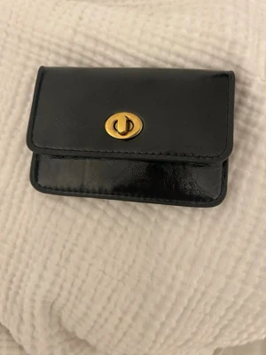 Svart läderplånbok med gulddetalj - Stilren svart plånbok i glansigt läder med en snygg guldfärgad vridknapp framtill. Plånboken har en kompakt rektangulär form och ett extra fack på baksidan för kort eller kvitton. Perfekt för dig som gillar klassisk och enkel design.