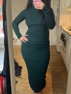 Mörkgrön ribbad långklänning - Snygg mörkgrön långklänning med ribbad struktur och lång ärm. Klänningen har en hög hals och figurnära passform som framhäver siluetten. Perfekt för dig som gillar stilrena och trendiga plagg.