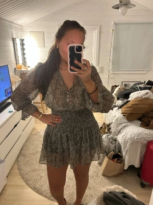 Leopardmönstrad kort klänning från Zara - Snygg kort klänning från Zara med leopardmönster i svart och beige. Klänningen har v-ringning, knappar framtill och långa, transparenta ärmar med volangdetaljer vid ärmslut och kjol. Tillverkad i ett lätt och luftigt syntetmaterial.