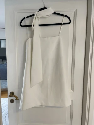Vit kort klänning med knytband - Jättesnygg elegant vit kort klänning som är axellös och ett coolt knytband runt halsen. Klänningen har en enkel och stilren design som känns modern och fräsch. Perfekt för dig som vill sticka ut med en unik detalj. Materialet känns slätt, men en av öglorna till knappen för bandet runt halsen behövs fixas. Aldrig använt. Är lite större i storlek.