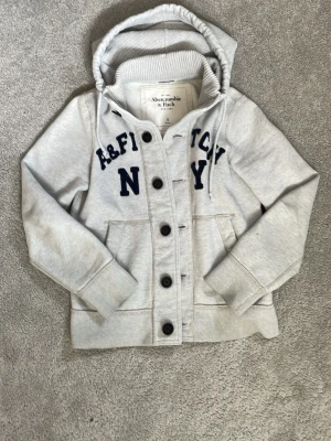 Grå hoodie från Abercrombie & Fitch - Snygg grå hoodie från Abercrombie & Fitch med knappar framtill och broderad text i mörkblått. Hoodien har huva med dragsko, två stora fickor och ribbade muddar. Perfekt för dig som gillar klassisk college-stil och mjuka material.